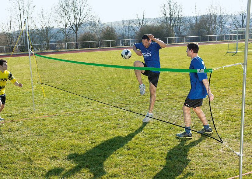 3 players playing soccer tennis, one player kicking ball with knee, outside picture