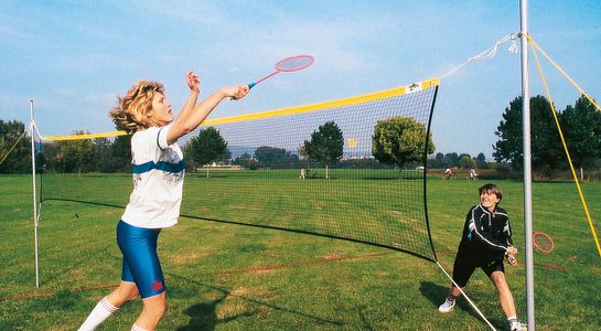 Badminton Net for leisure, with yellow headband, 2 female players, action scene, one jumping to hit the ball with racket, playing on green gras, outdoors